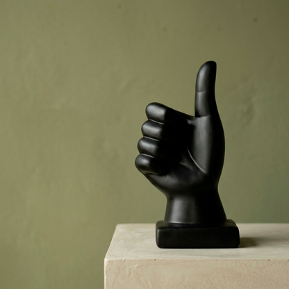 Escultura decorativa de mano con pulgar arriba, gesto “like”, en cerámica negra mate sobre base cuadrada, en ambiente de pared verde
