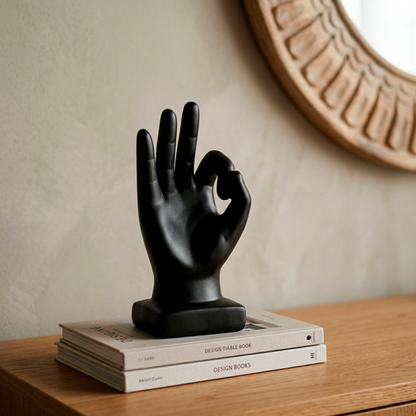Escultura decorativa de mano con gesto OK en cerámica negra mate, sobre libros de diseño junto a un espejo de madera