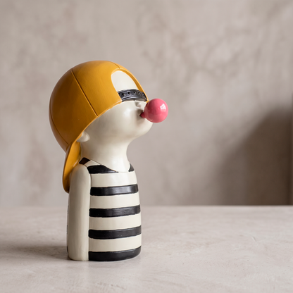 Figura decorativa de niño con gorra mostaza y camiseta a rayas soplando un globo de chicle rosado, en resina marfil, sobre mesa neutra